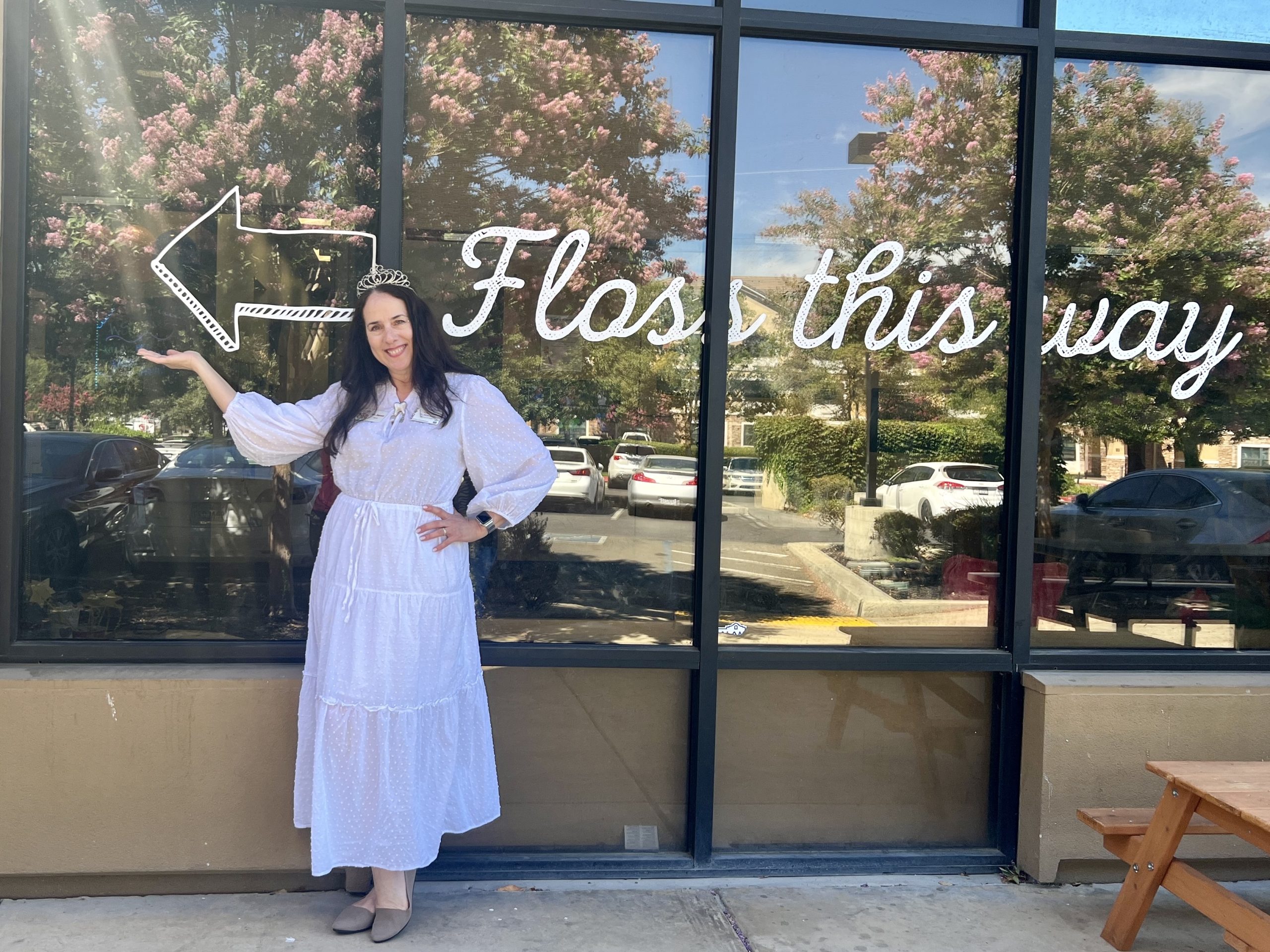 Team member at Kids Care Dental Tracy dressed as the tooth fairy standing in front of exterior signage that reads 'Floss This Way'