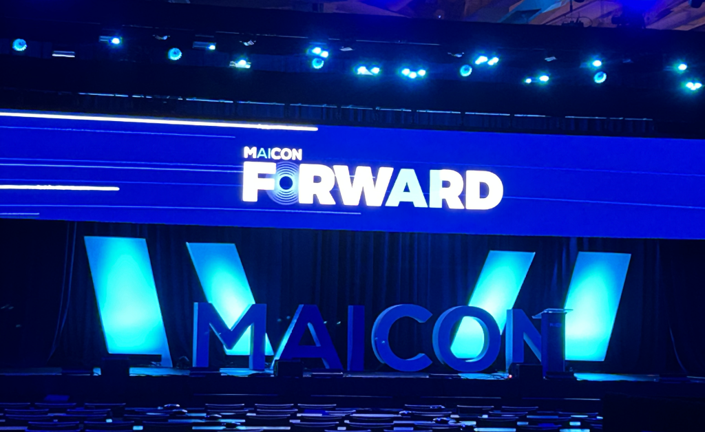 MAICON main stage before a session, with the “MAICON Forward” theme lit on a blue screen above rows of empty seats